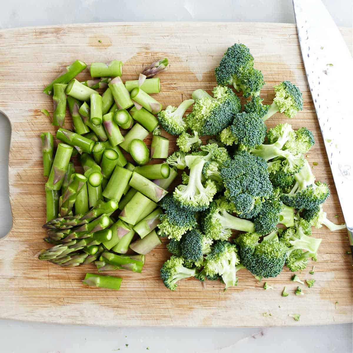 Chopped broccoli florets and asparagus on a cutting board with a knife.
