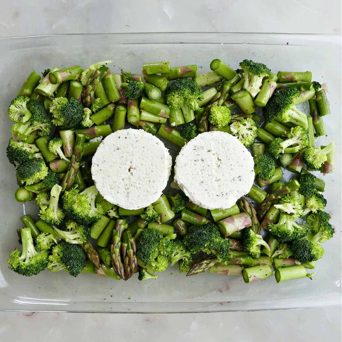 Broccoli and asparagus in a large glass baking dish topped with two wheels of cheese.