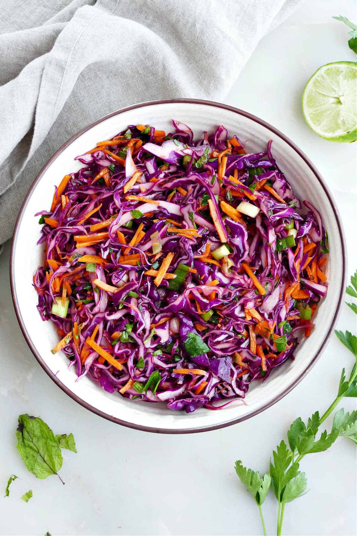 Cabbage carrot slaw in a large bowl next to a kitchen towel and lime half.