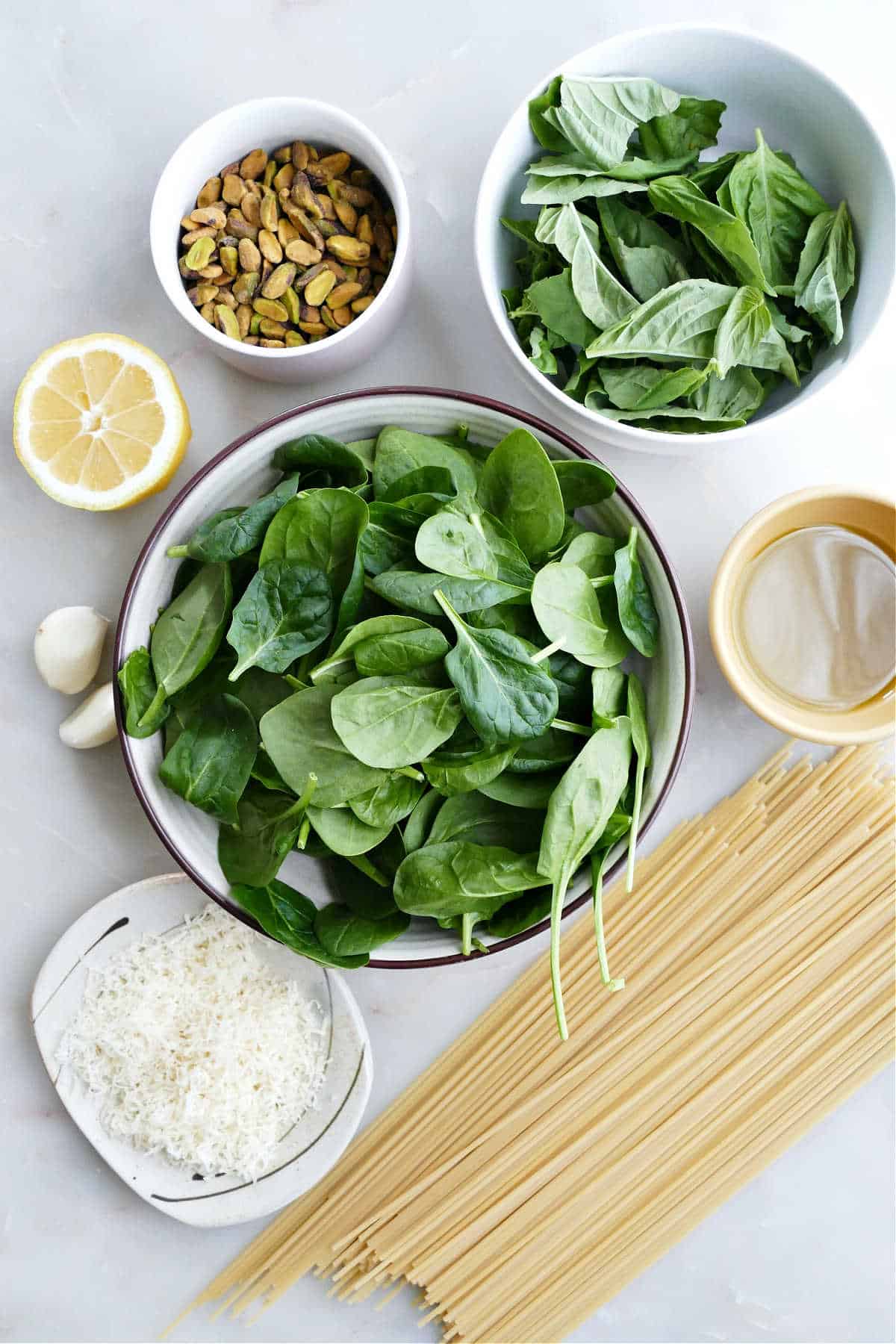 Pistachios, lemon, spinach, basil, spaghetti, olive oil, and cheese on a counter.