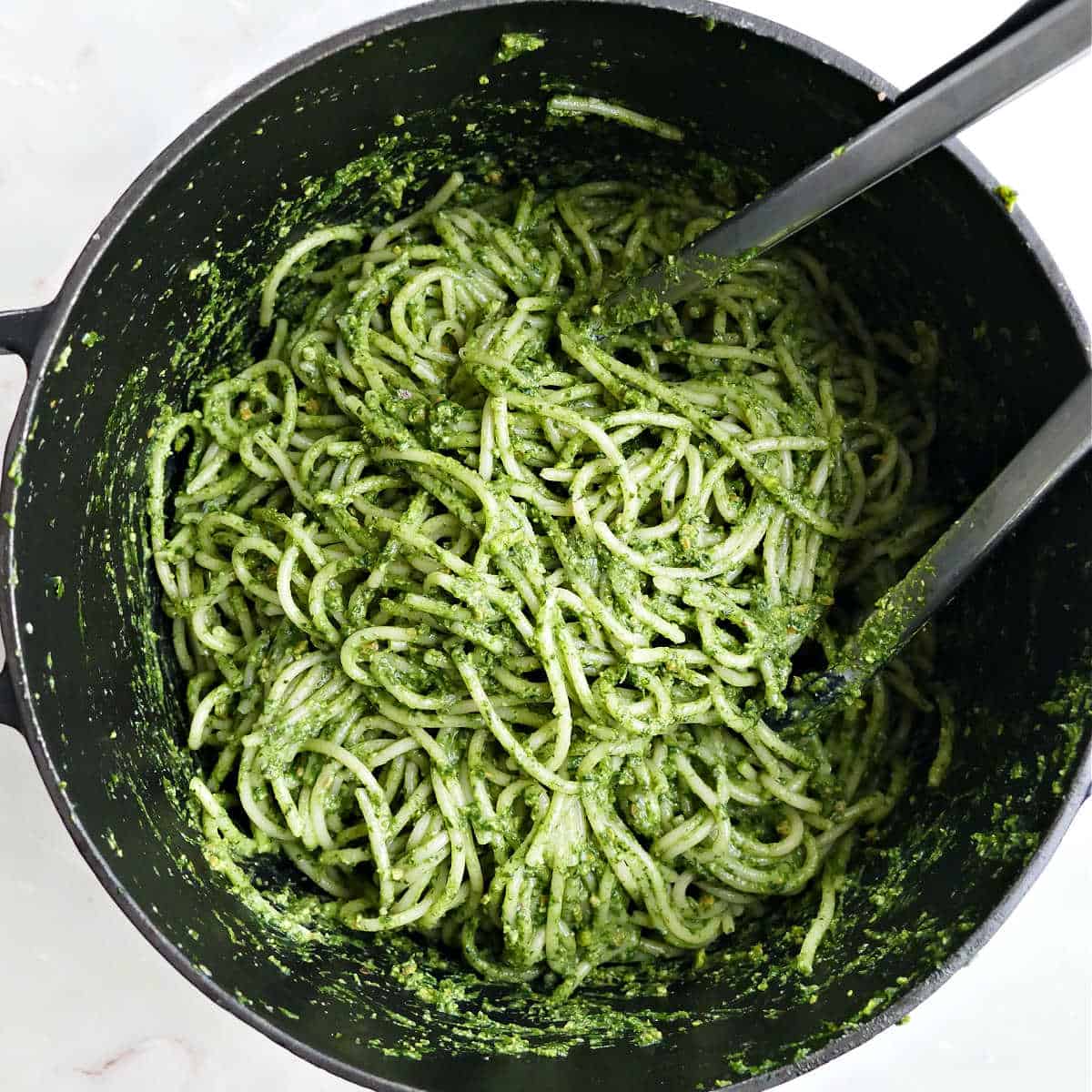 Spaghetti tossed with green pasta sauce in a large pot with tongs.