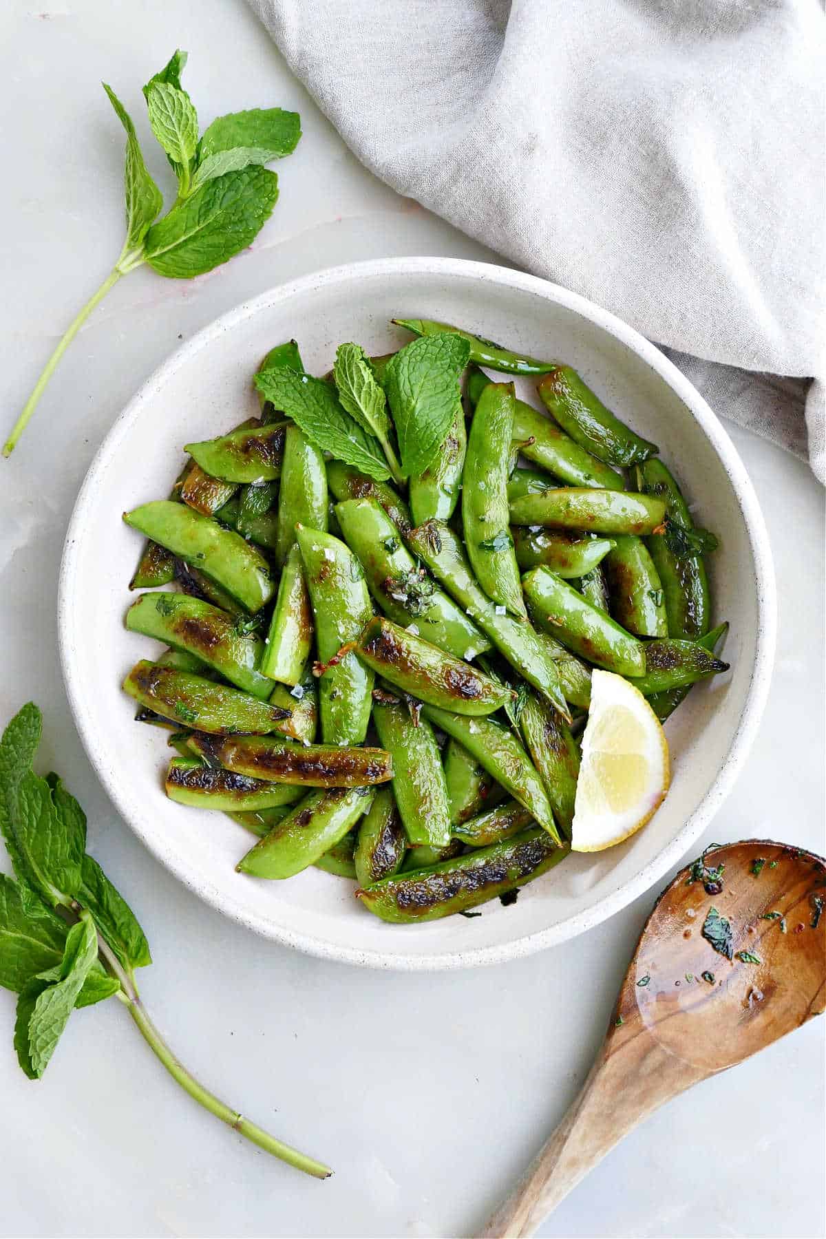 Sauteed snap peas in a large white bowl with a lemon next to garnish and a wooden spoon.