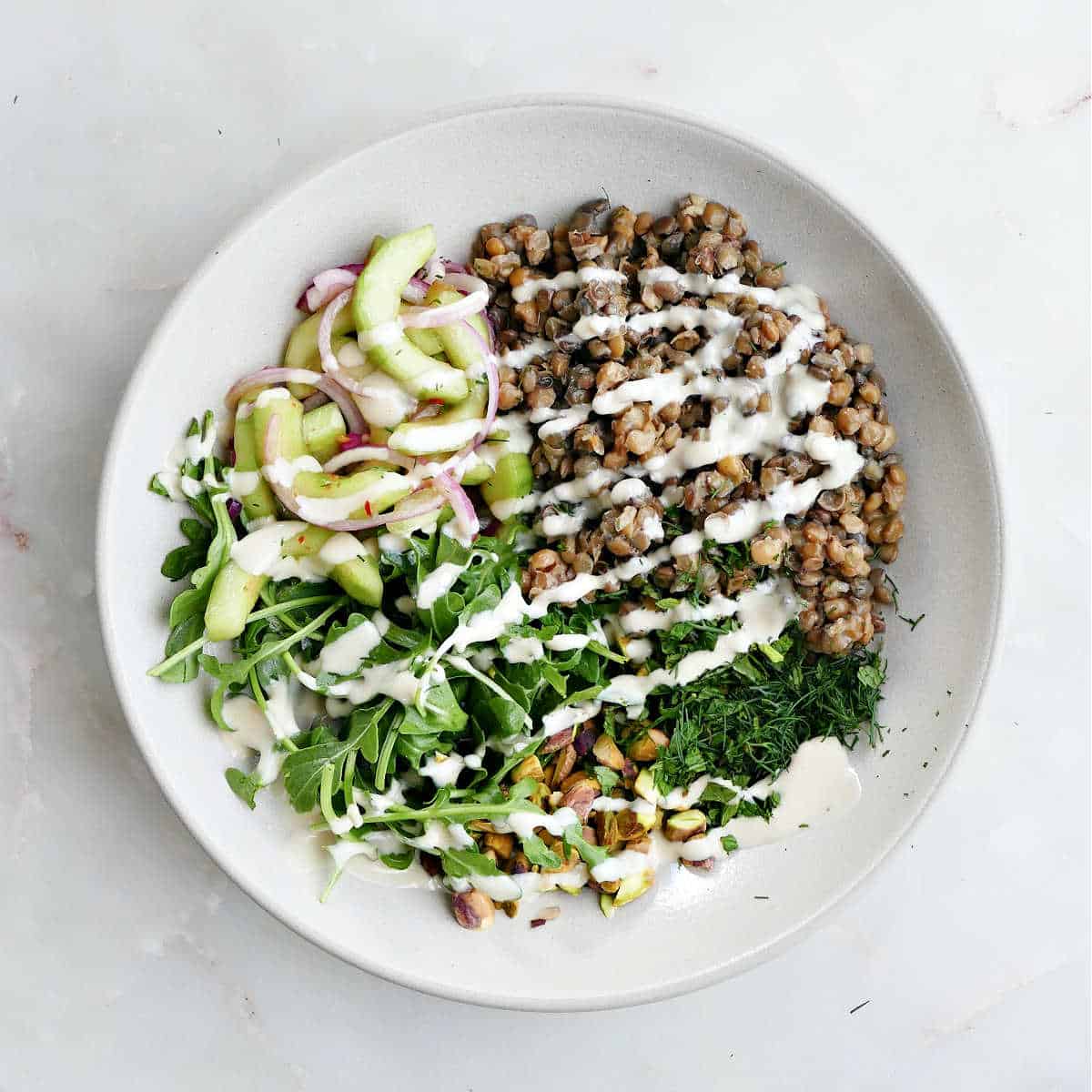 A lentil bowl with cucumber, onions, nuts, and salad in a large bowl with dressing drizzled over top.