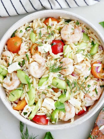 A bowl of shrimp orzo salad topped with garnish next to a wooden spoon.