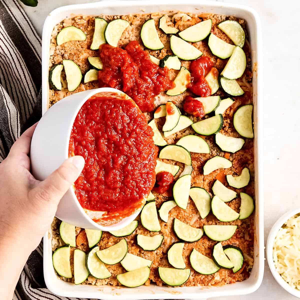 Woman adding marinara sauce on top of zucchini pieces and casserole filling in a dish.