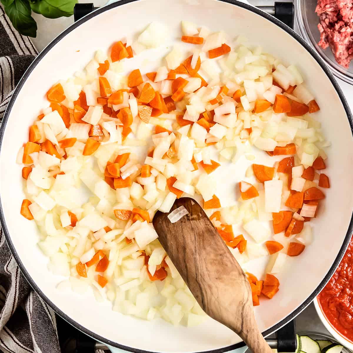 Carrots and onions cooking in olive oil in a skillet being stirred with a wooden spoon.