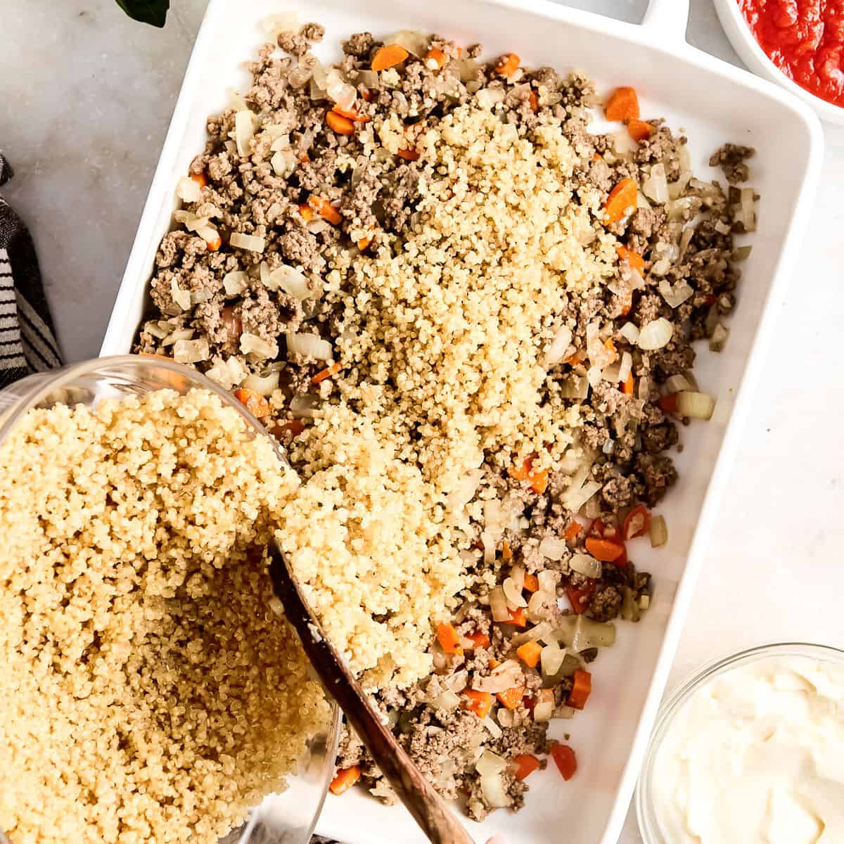 Woman adding cooked quinoa to a casserole dish with beef and veggie mixture.