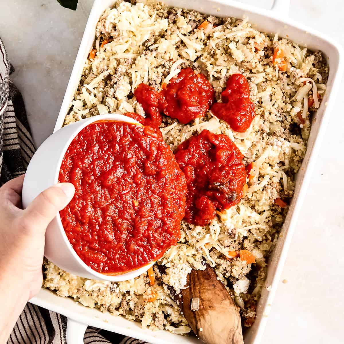 Woman pouring marinara sauce in a casserole dish with beef, veggies, and quinoa.