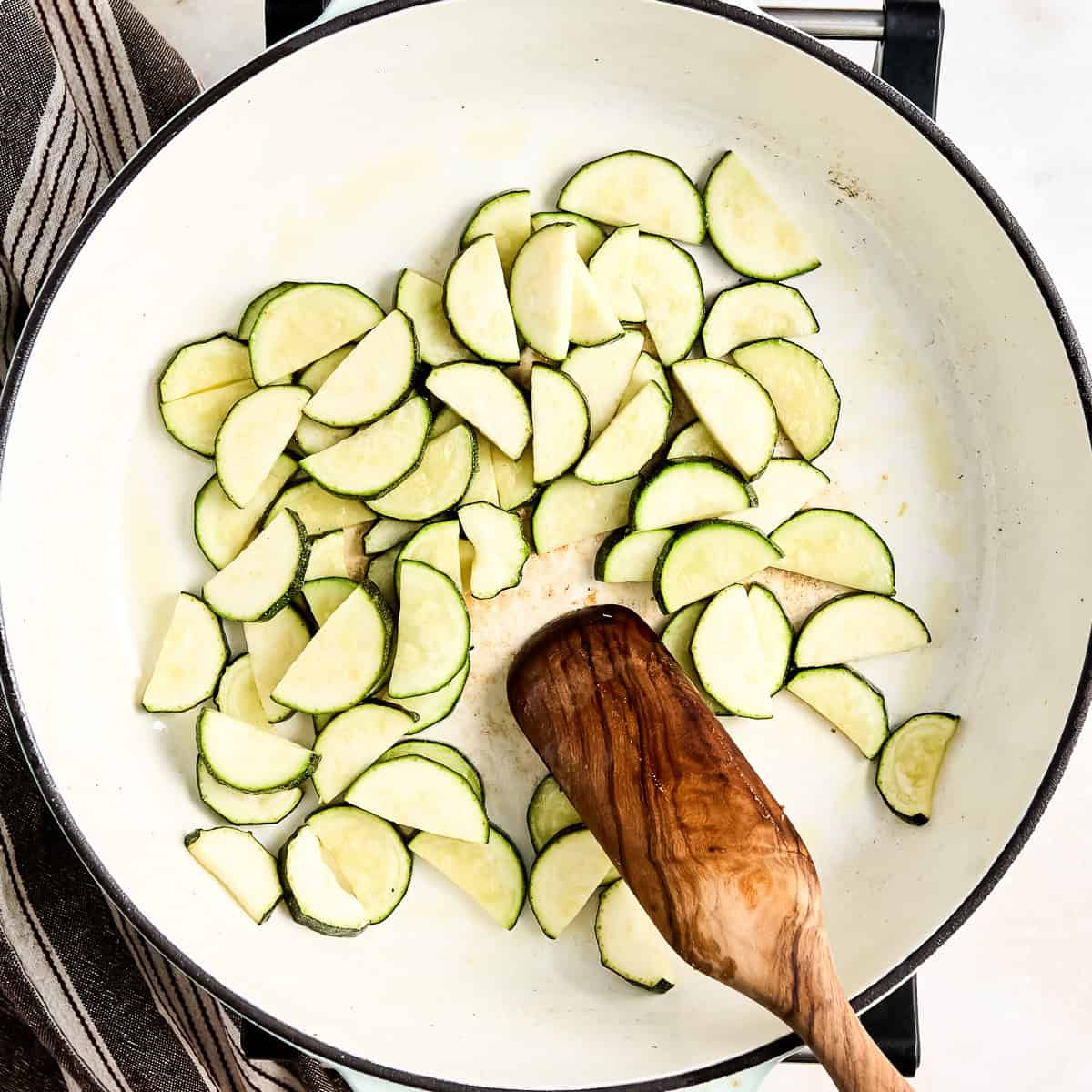 Zucchini pieces cooking in olive oil in a large skillet on a hot plate.