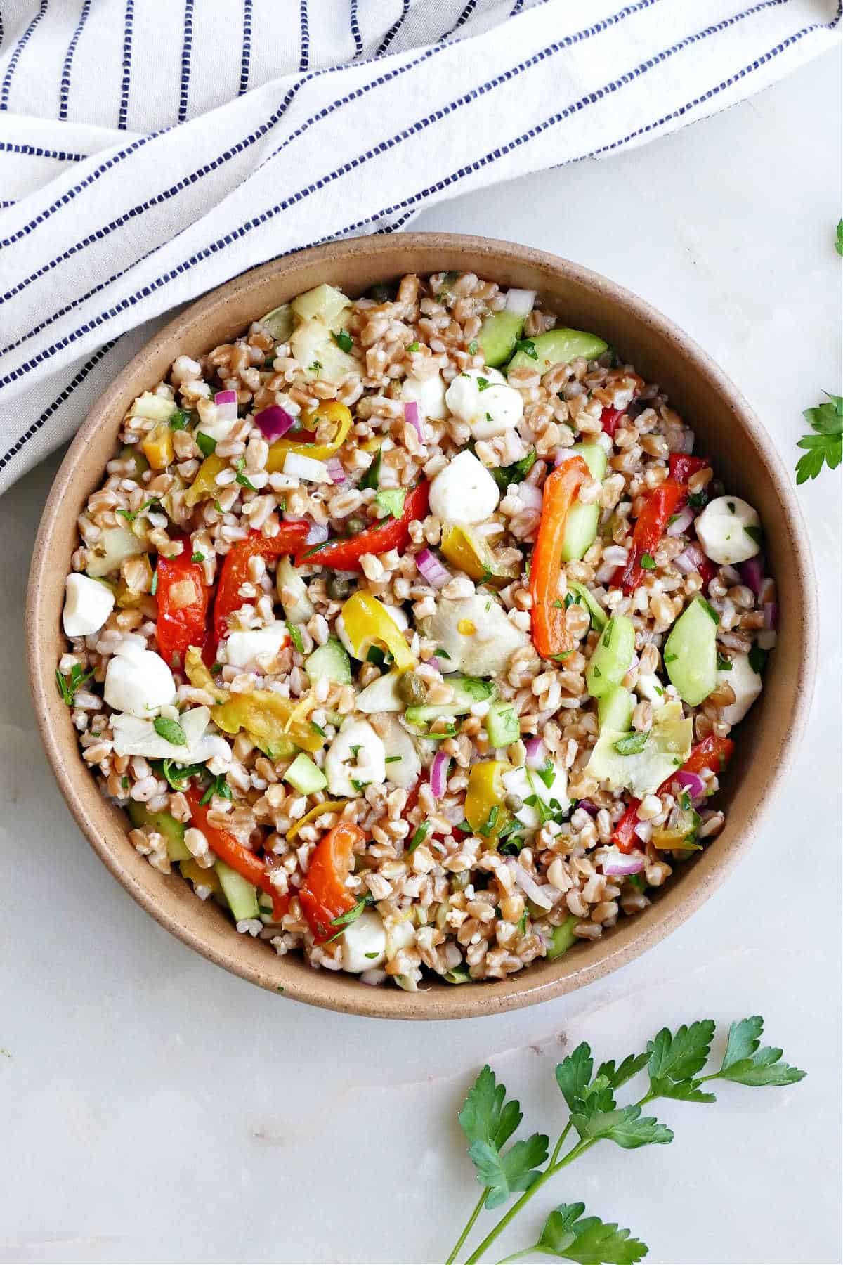 Cold Italian farro salad in a brown serving bowl next to a kitchen towel and parsley.