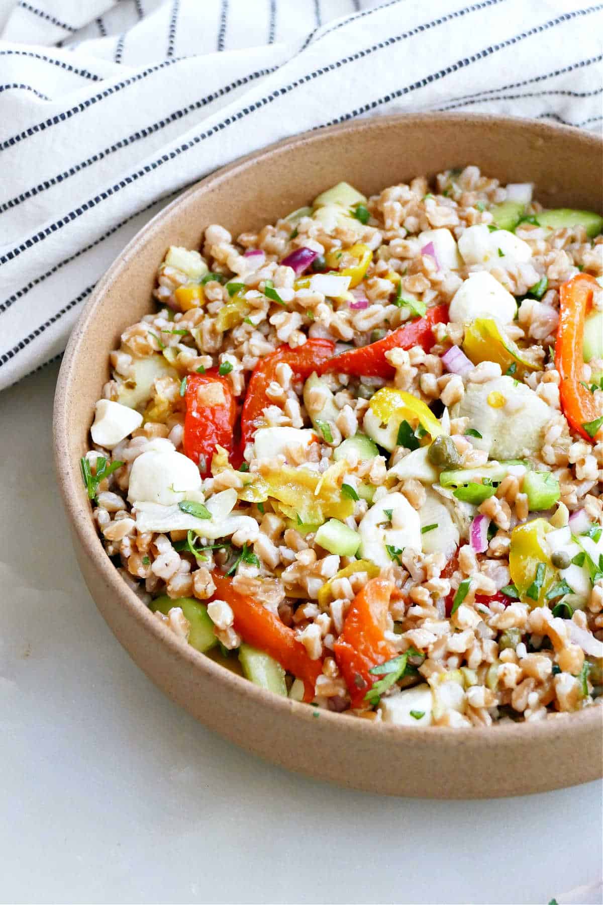 Cold Italian farro salad in a large brown serving bowl next to a striped kitchen towel.