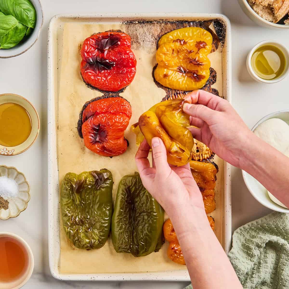 Woman peeling skin from a roasted yellow bell pepper over baking sheet with others.