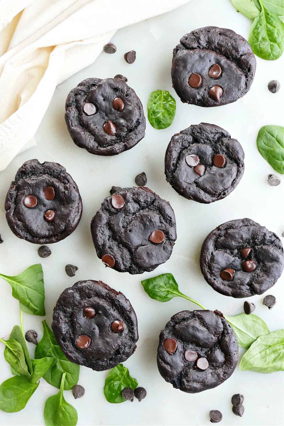 8 spinach chocolate muffins next to chocolate chips and baby spinach leaves on a counter.