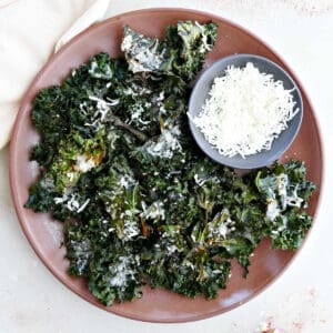 Crispy parmesan kale chips on a platter with a bowl of cheese.