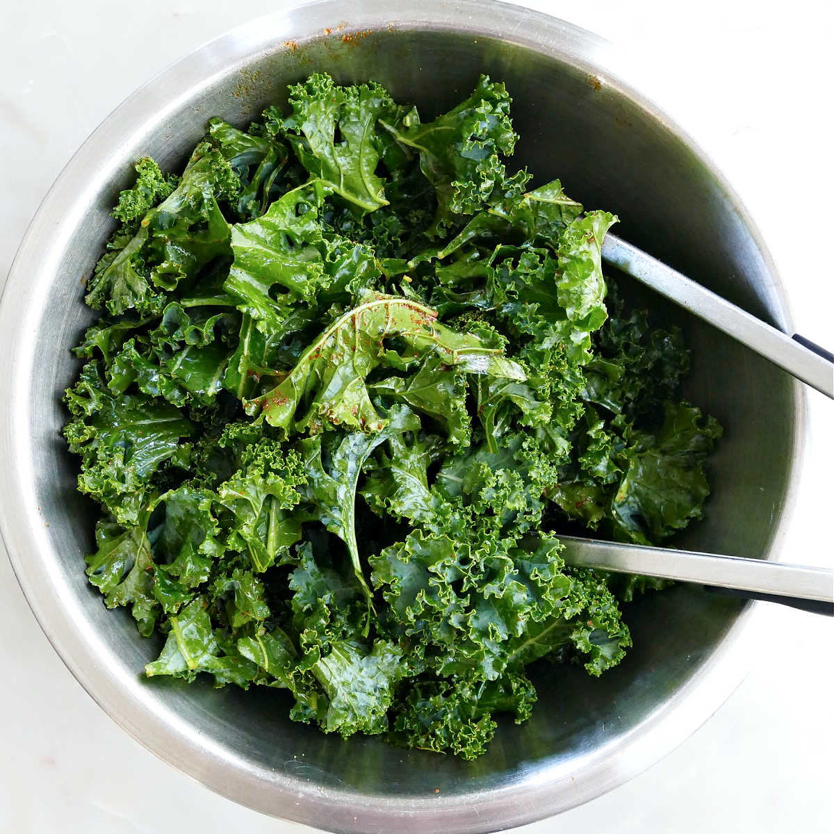 Torn kale pieces being tossed in oil and spices in a bowl with tongs.