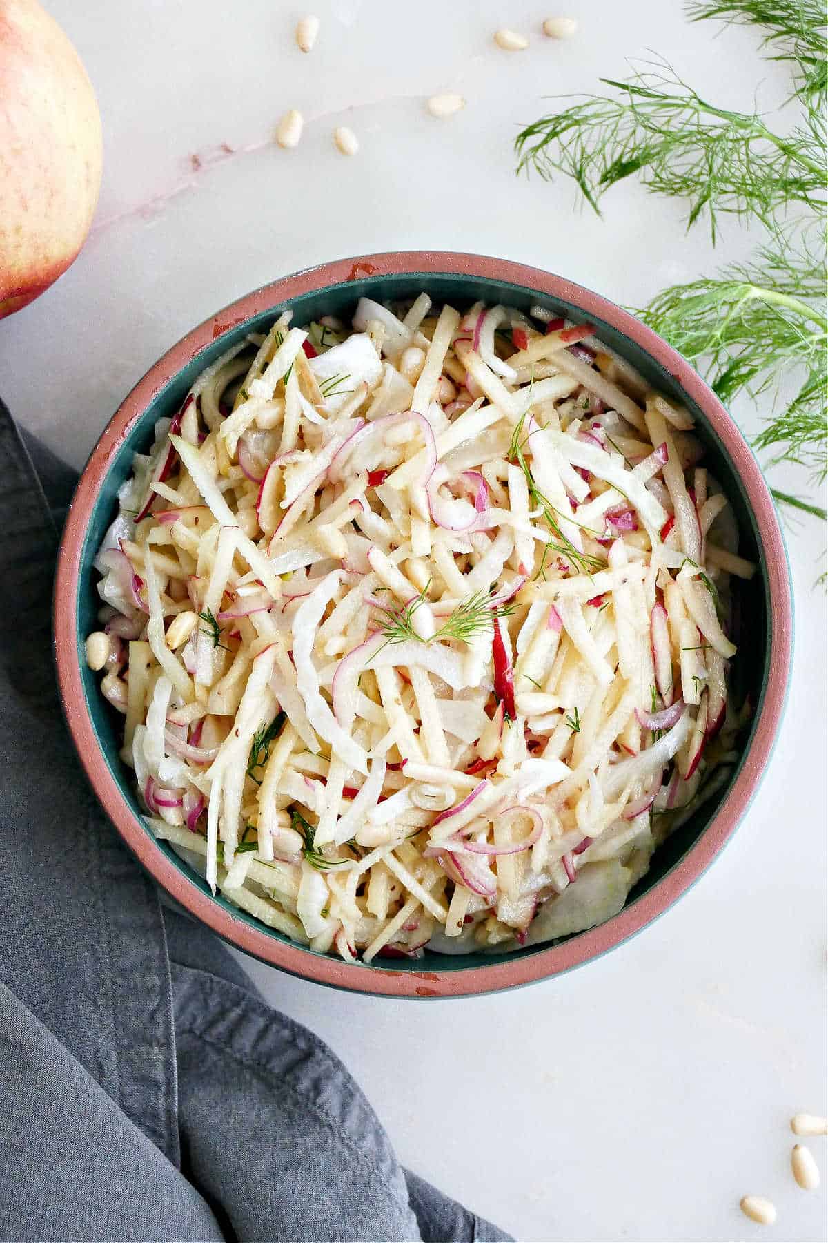 Fennel apple salad in a serving bowl next to garnishes and a napkin on a counter.