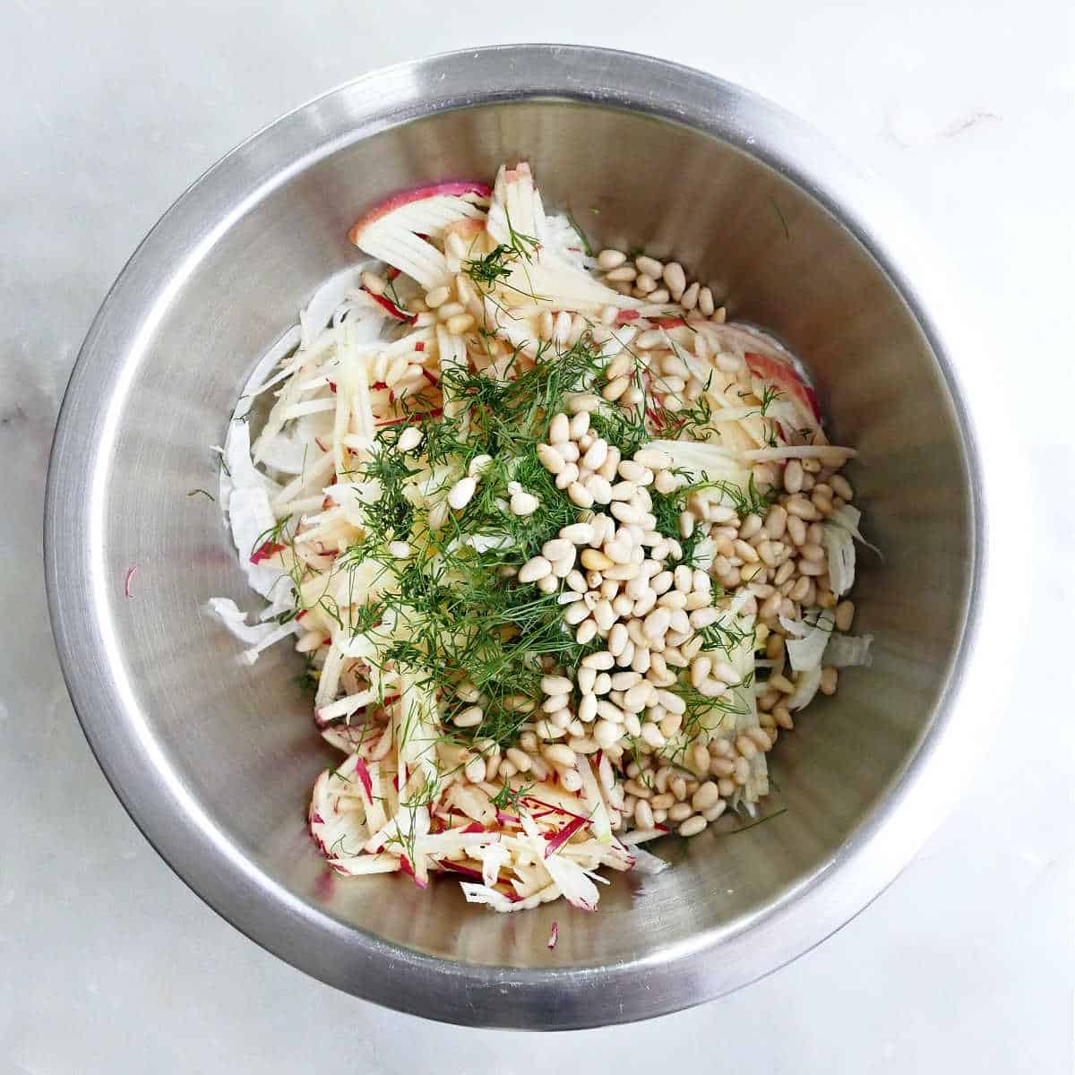 Fresh herbs, apples, fennel, and pine nuts in a bowl.