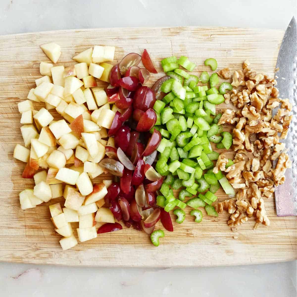 Chopped grapes, celery, apples, and nuts on a wooden cutting board with a knife.