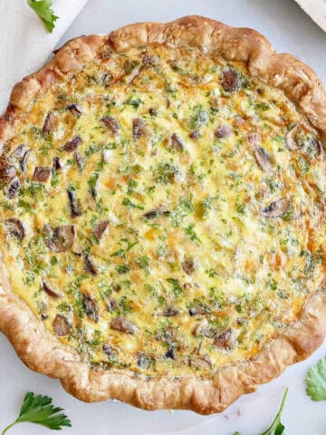 Loaded vegetarian quiche next to gruyere cheese, parsley, eggs, and napkin.