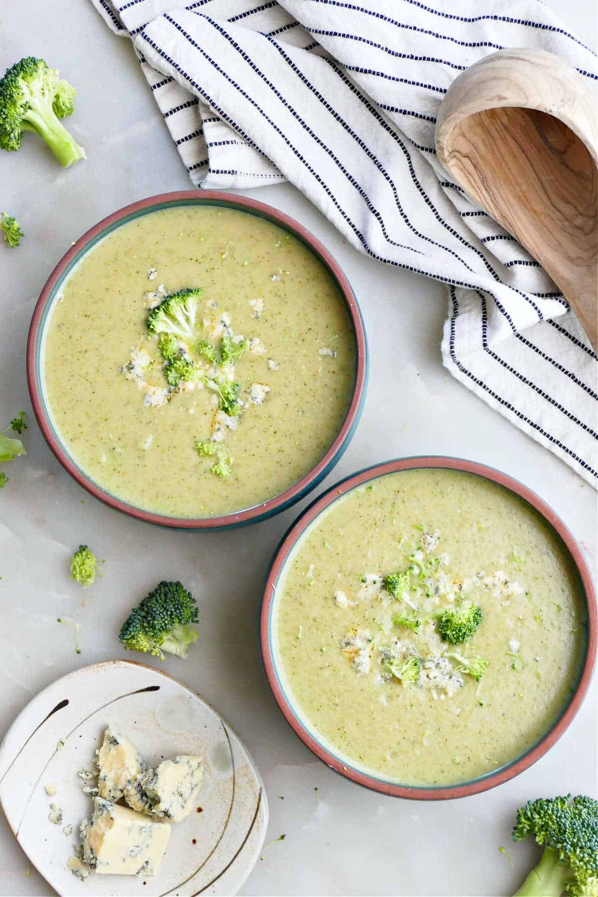 Two bowls of broccoli and Stilton soup next to toppings.