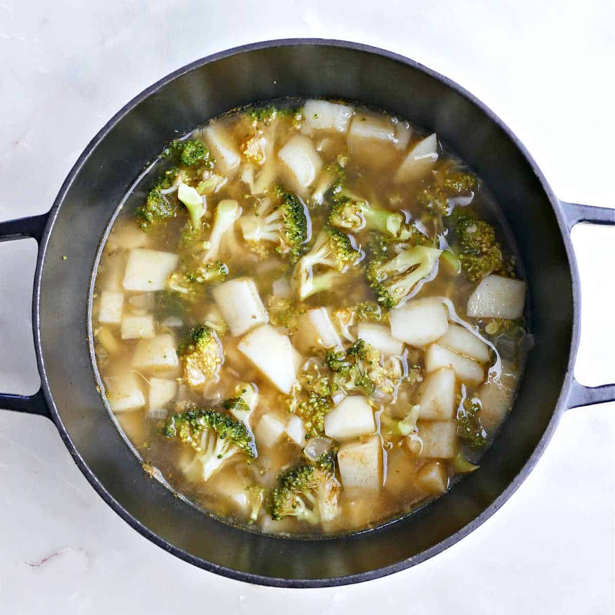 Broth added to ingredients cooking for broccoli and Stilton soup in a pot.