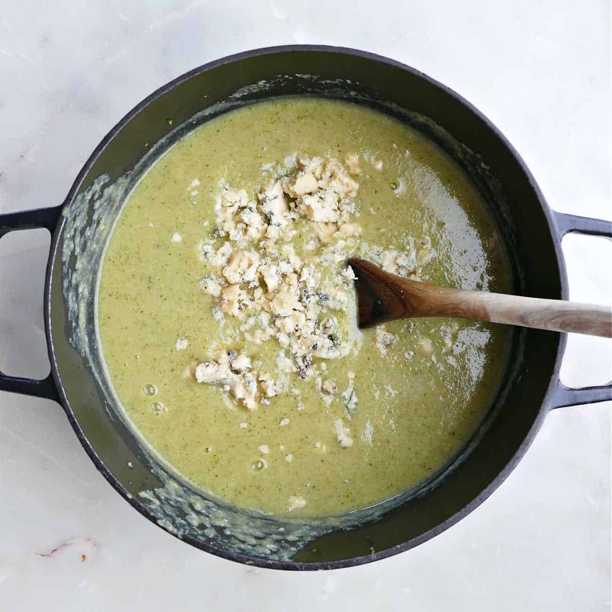 Blended broccoli soup in a pot with blue cheese crumbles and a spoon.