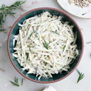 Celeriac coleslaw in a bowl on a table surrounded by garnishes.