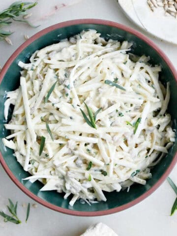 Celeriac coleslaw in a bowl on a table surrounded by garnishes.