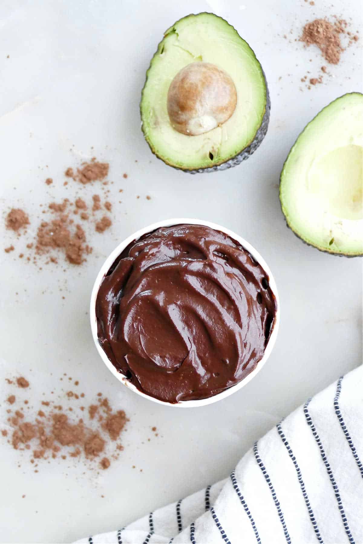 Healthy chocolate frosting with avocado in a cup next to avocados and a napkin.