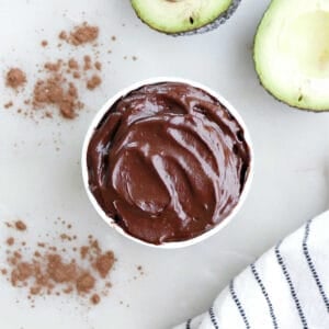 Healthy chocolate frosting with avocado in a cup next to avocados and a napkin.