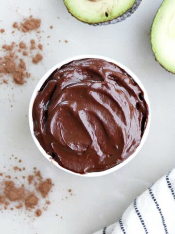 Healthy chocolate frosting with avocado in a cup next to avocados and a napkin.