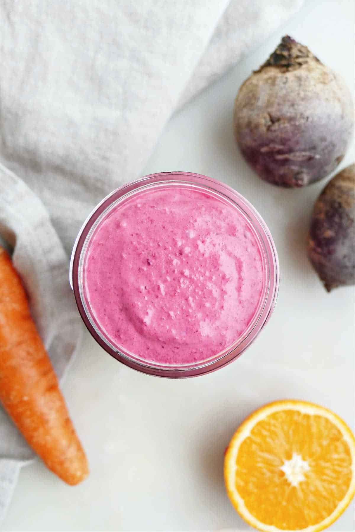 Beet carrot smoothie in a glass on a counter surrounded by ingredients.