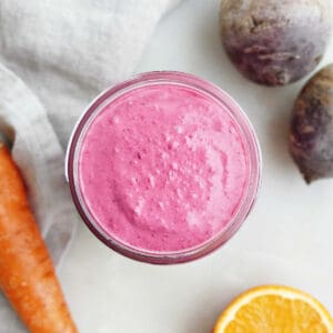 Beet carrot smoothie in a glass on a counter surrounded by ingredients.