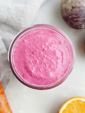 Beet carrot smoothie in a glass on a counter surrounded by ingredients.