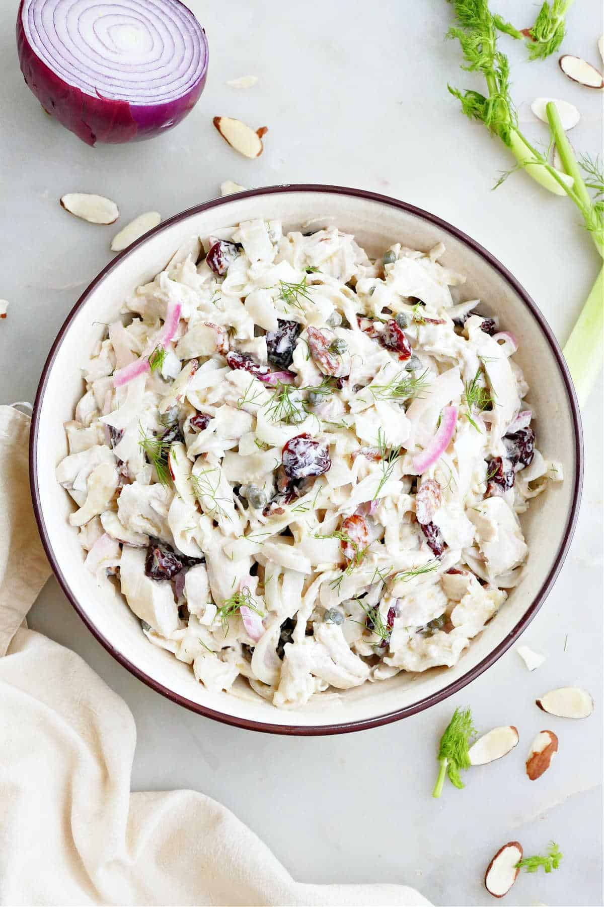 Chicken fennel salad in a serving bowl surrounded by ingredients and a napkin.