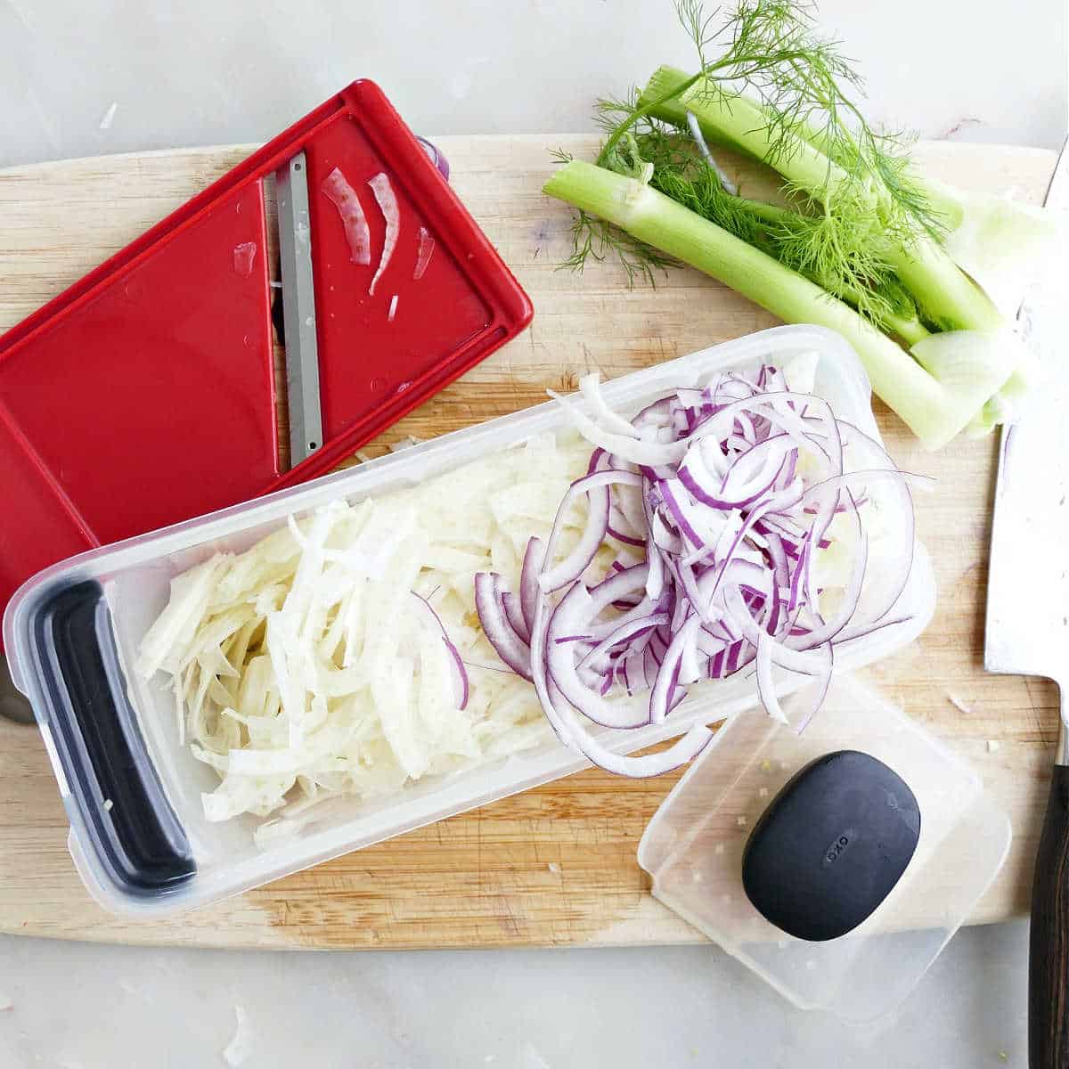 Shaved fennel and red onion in a mandolin on a cutting board.