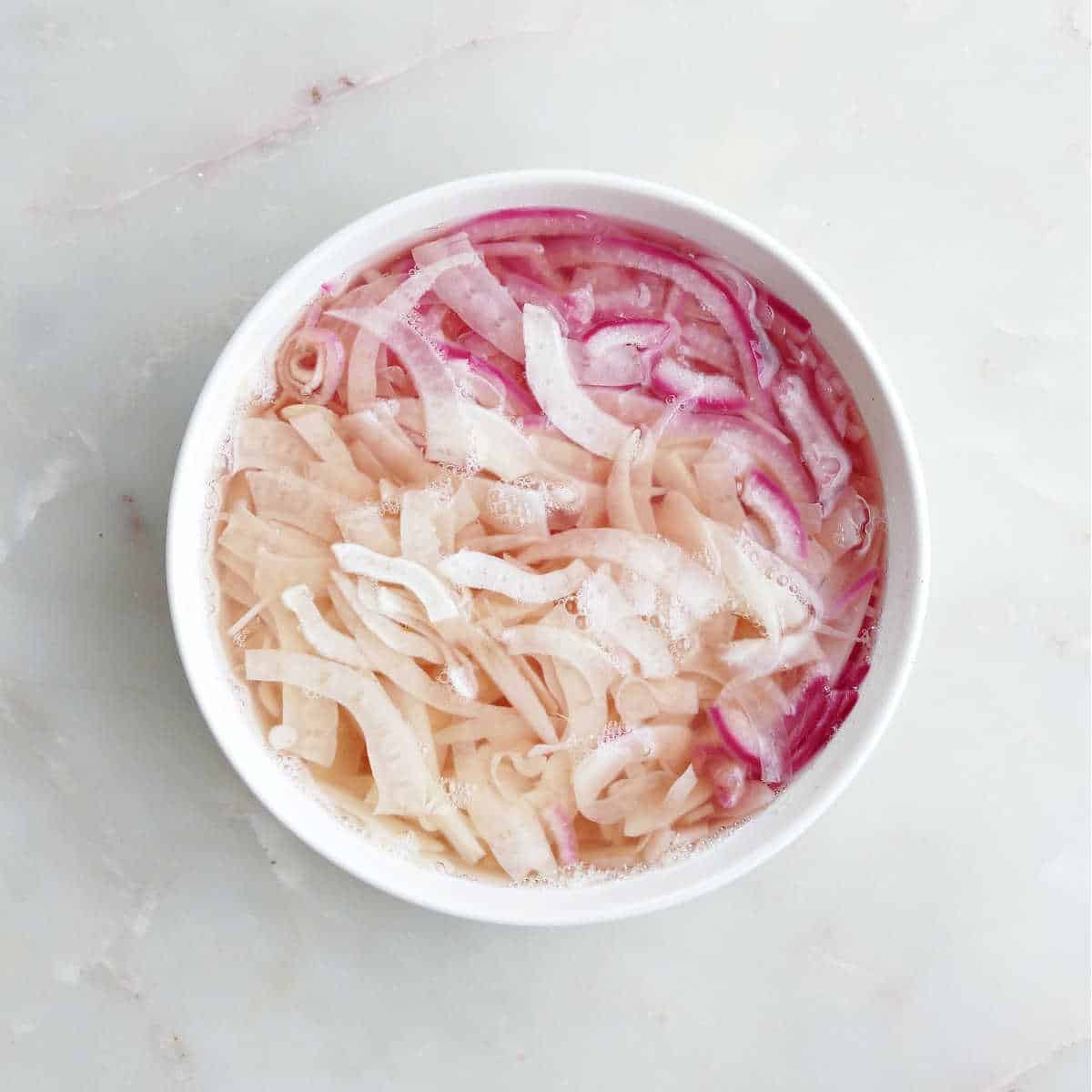 Shaved fennel and red onion in pickling brine in a bowl.