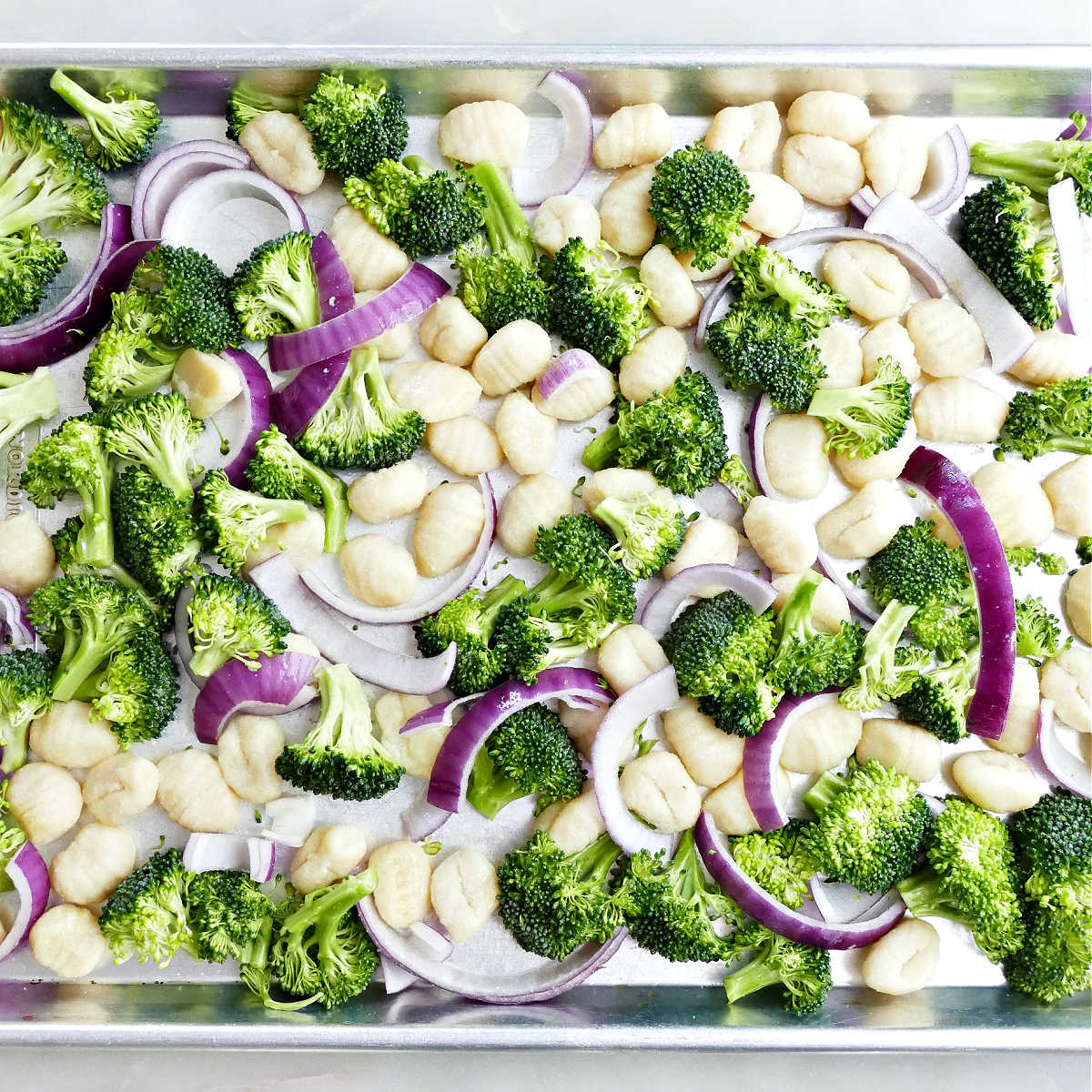 Gnocchi, broccoli, and red onion on a half sheet pan before roasting.