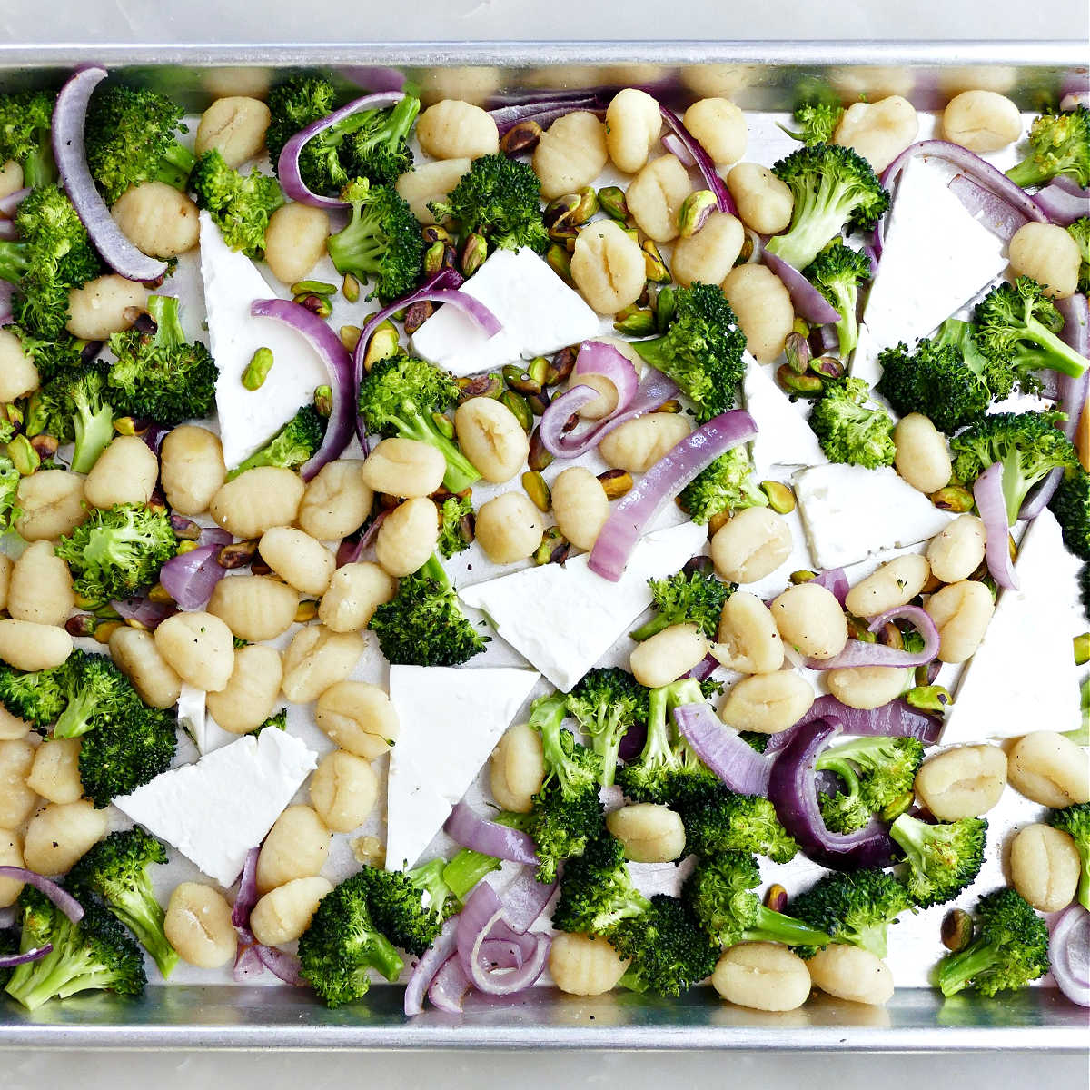 Planks of feta cheese added to a sheet pan of gnocchi, red onion, and broccoli.