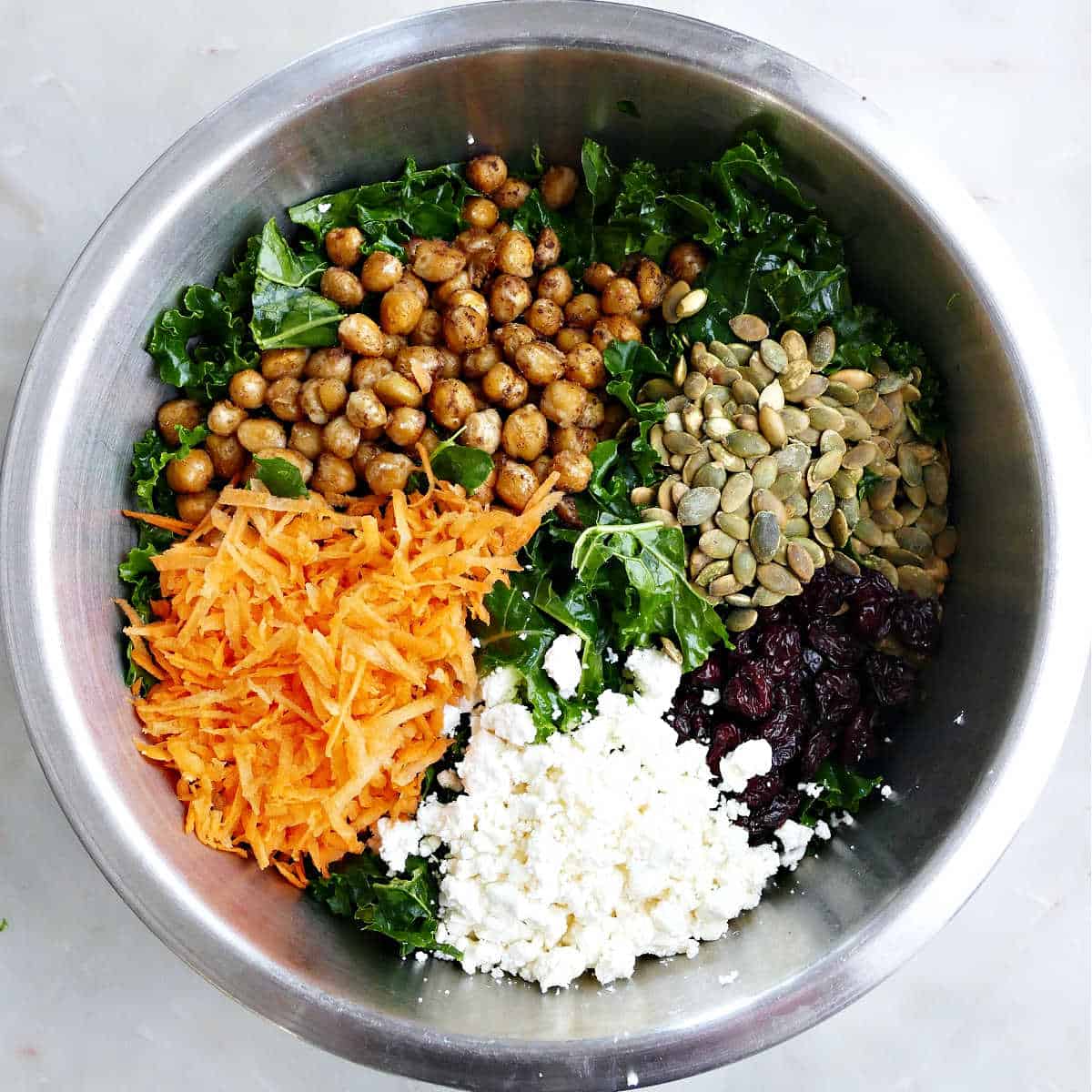 Kale, chickpeas, carrots, pumpkin seeds, feta, and dried cherries in a mixing bowl.