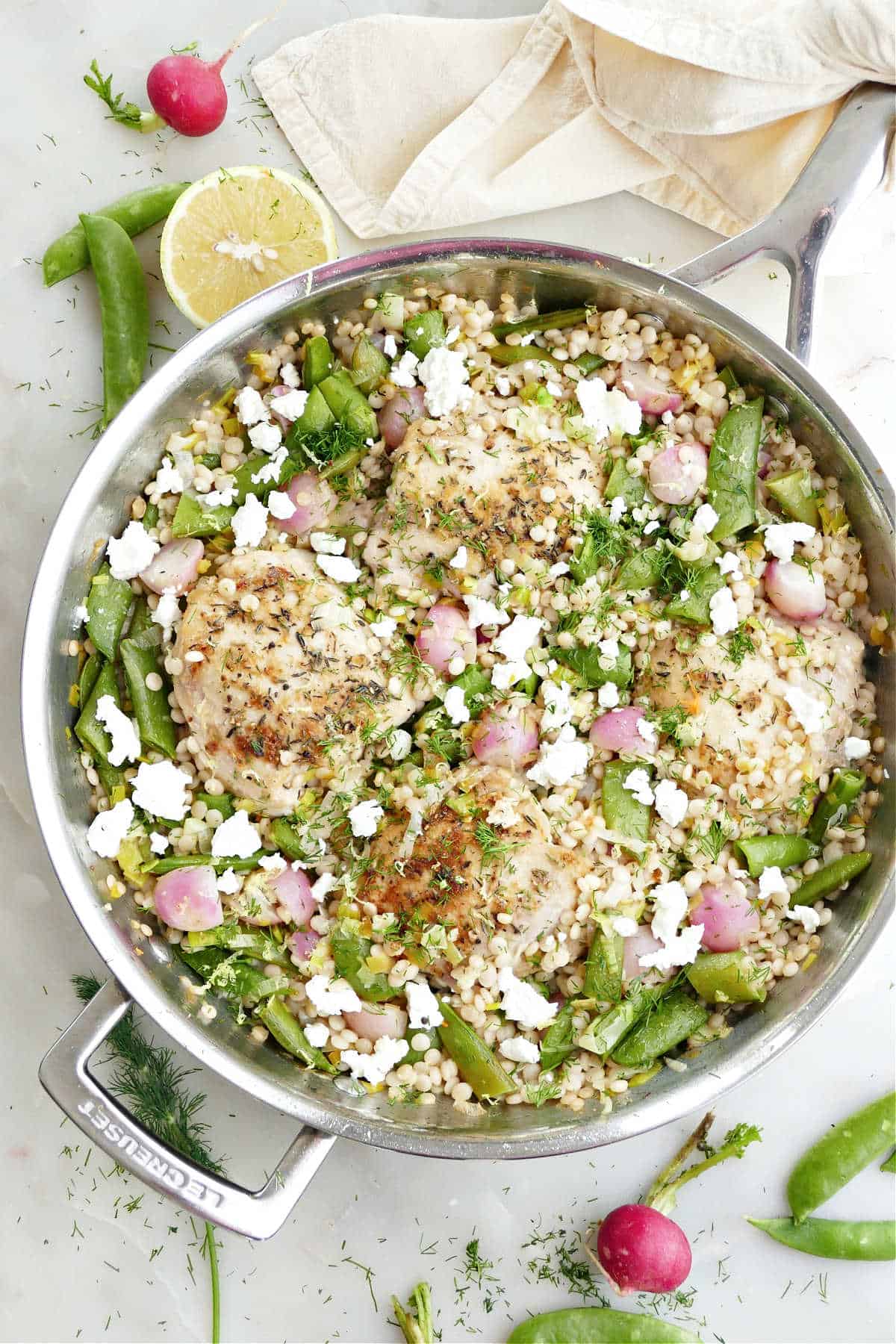 Large skillet with chicken thighs, couscous, and vegetables next to ingredients.