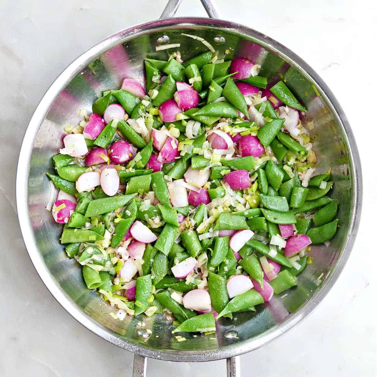 Leeks, radishes, snap peas, and garlic cooking in oil in a skillet.