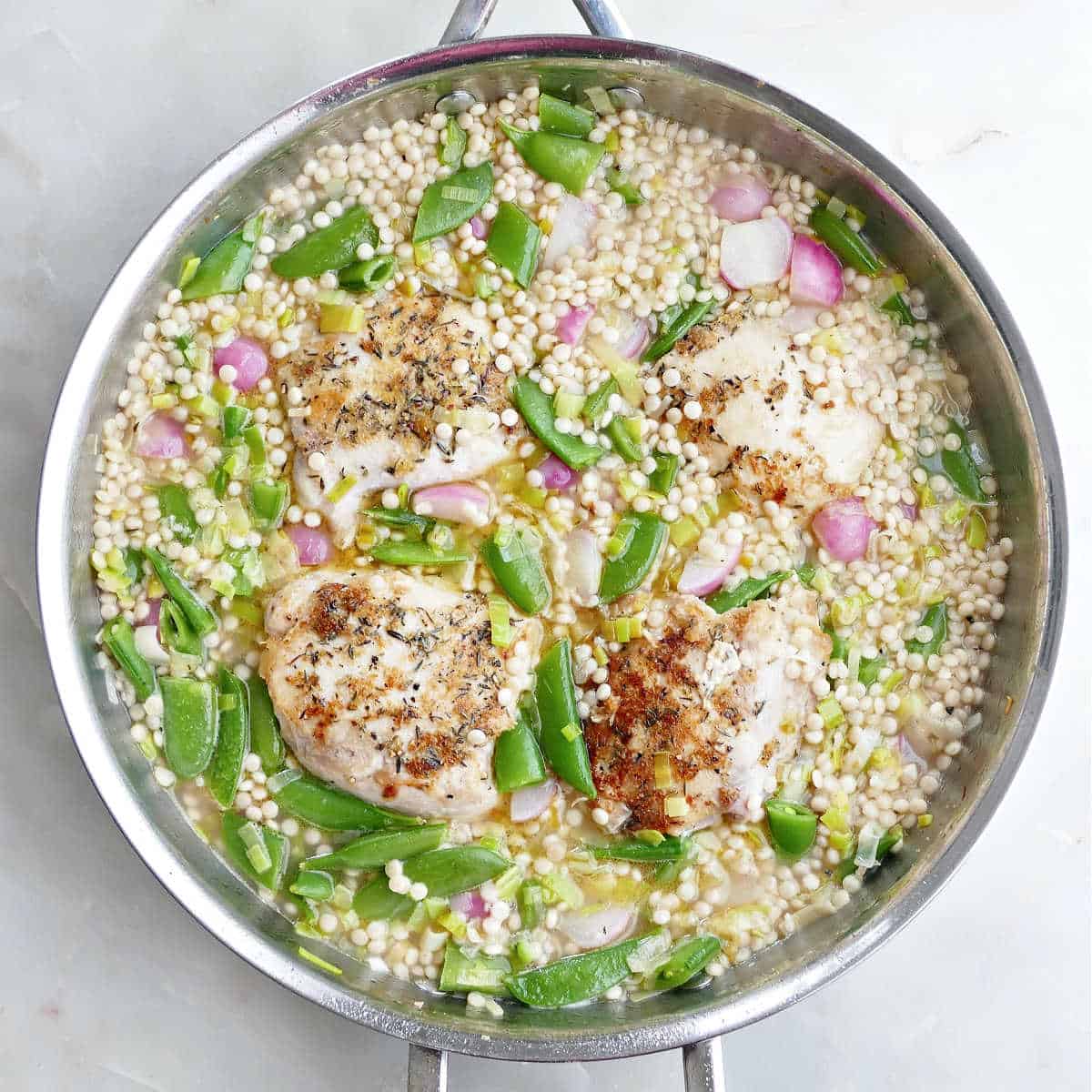 Broth, couscous, vegetables, and chicken thighs in a skillet before baking.
