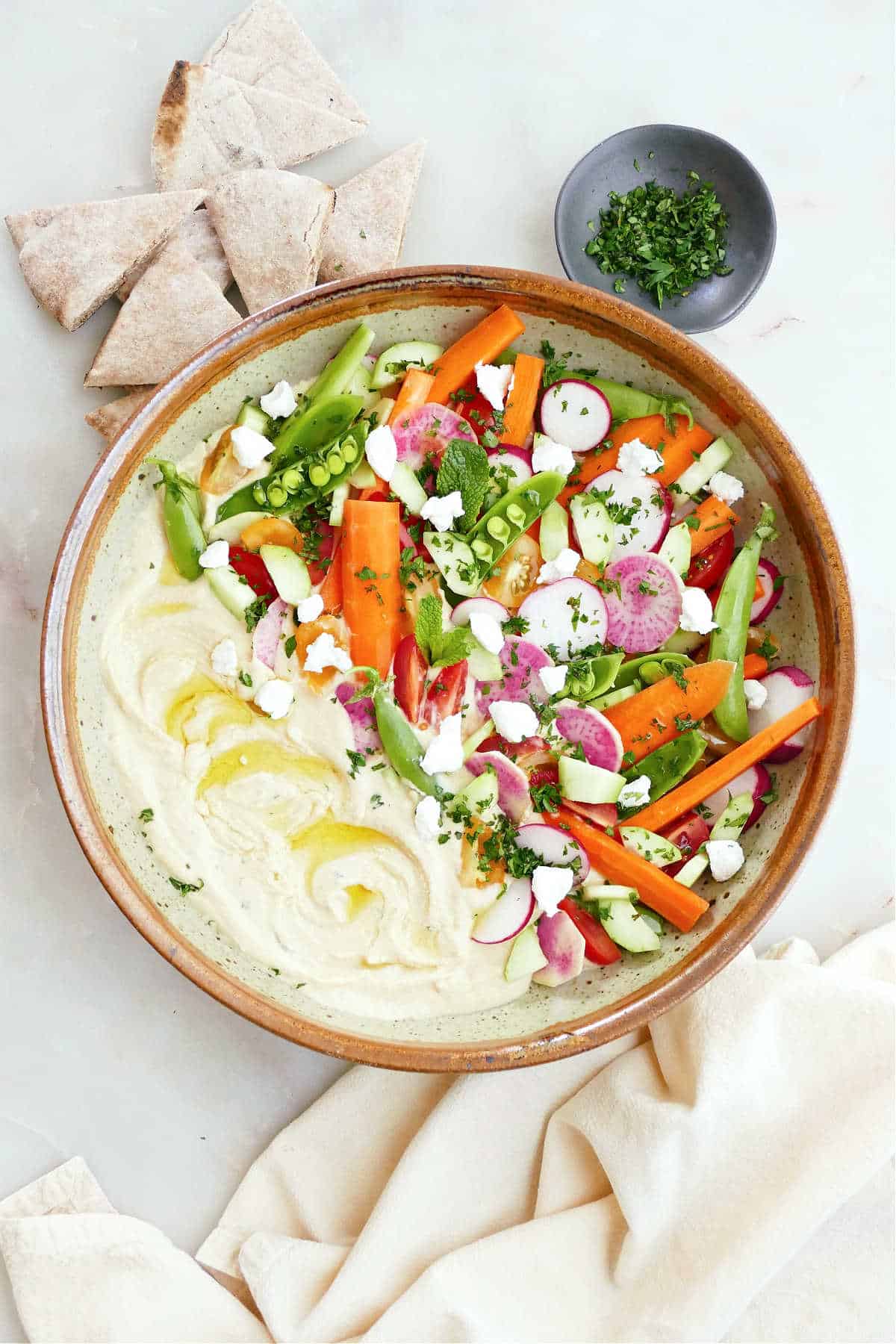 Large bowl with hummus topped with colorful vegetables next to pita, a napkin, and herbs.