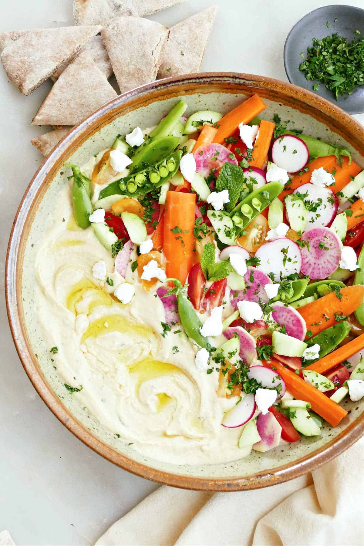 Serving bowl with hummus, carrots, radishes, snap peas, tomatoes, herbs, and goat cheese.