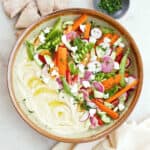Large bowl with hummus topped with colorful vegetables next to pita, a napkin, and herbs.