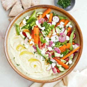 Large bowl with hummus topped with colorful vegetables next to pita, a napkin, and herbs.