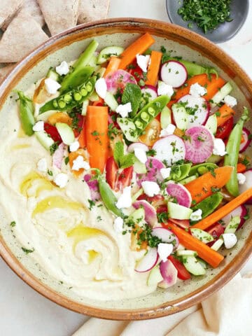 Large bowl with hummus topped with colorful vegetables next to pita, a napkin, and herbs.