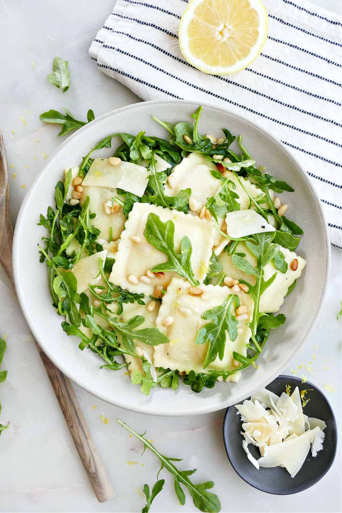 Ravioli pasta salad with arugula in a bowl surrounded by napkin, lemon, spoon, and cheese.
