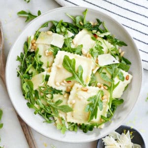 Ravioli pasta salad with arugula in a bowl surrounded by napkin, lemon, spoon, and cheese.